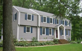 An exterior view of a two-story traditional home featuring light gray horizontal vinyl siding, white trim, and navy blue decorative shutters on the windows, set against a backdrop of lush green trees.