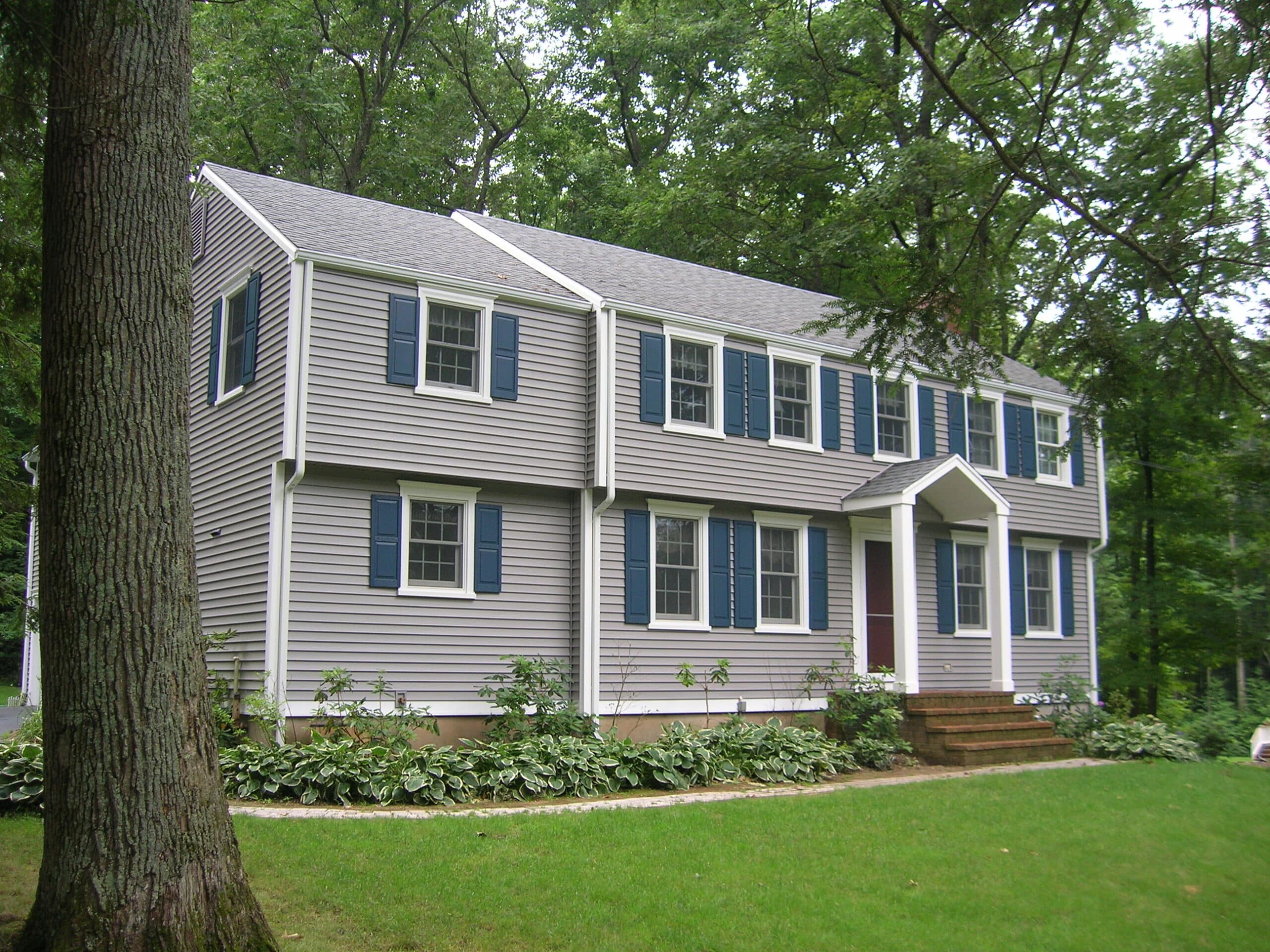 gray-vinyl-siding-home-with-blue-shutters An exterior view of a two-story traditional home featuring light gray horizontal vinyl siding, white trim, and navy blue decorative shutters on the windows, set against a backdrop of lush green trees.