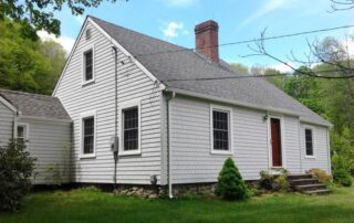 The exterior of a classic Cape Cod style house featuring white shingle siding, a red front door, a brick chimney, and a fieldstone foundation, surrounded by green grass and trees.