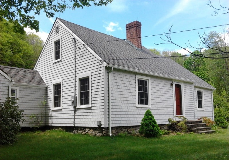white-shingle-cape-cod-home-exterior The exterior of a classic Cape Cod style house featuring white shingle siding, a red front door, a brick chimney, and a fieldstone foundation, surrounded by green grass and trees.