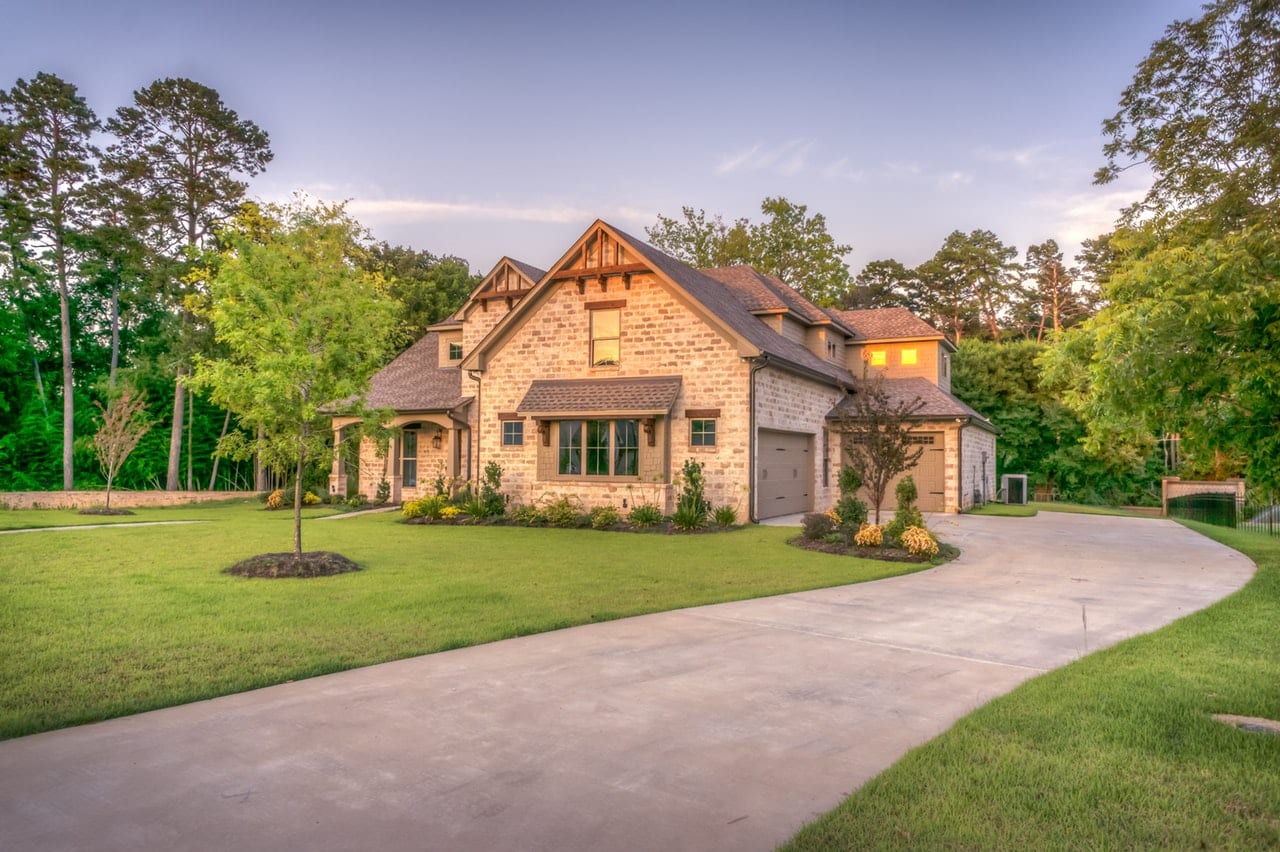 luxury-stone-exterior-home-with-curved-driveway A large, elegant stone house with multiple gables and a sprawling green lawn, featuring a long, curved concrete driveway leading to a side-entry garage under a soft sunset sky.