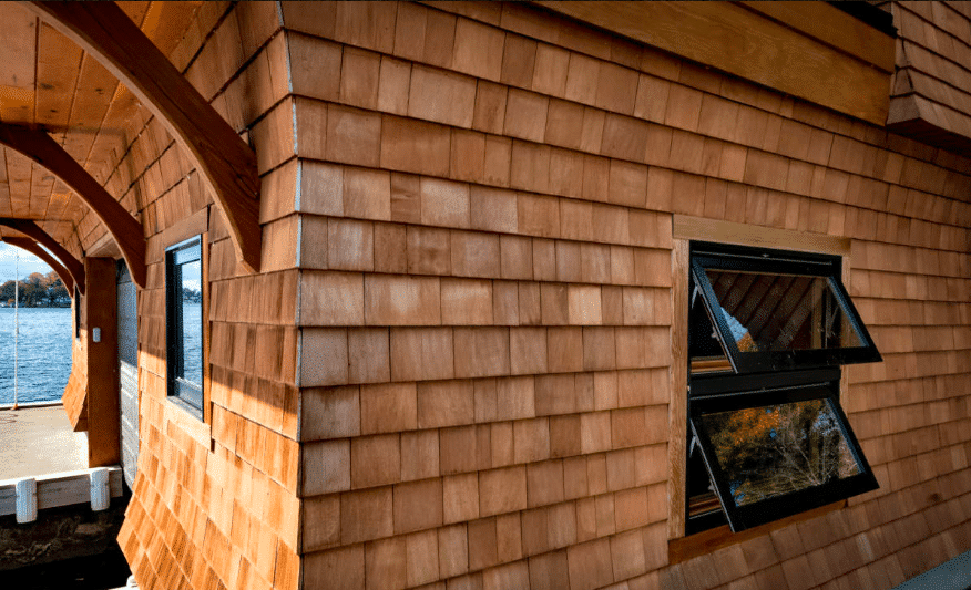 cedar-shingle-siding-with-awning-windows Exterior view of a lakeside building featuring warm-toned cedar shingle siding and black-framed awning windows propped open, with a view of a covered walkway and the water in the background.